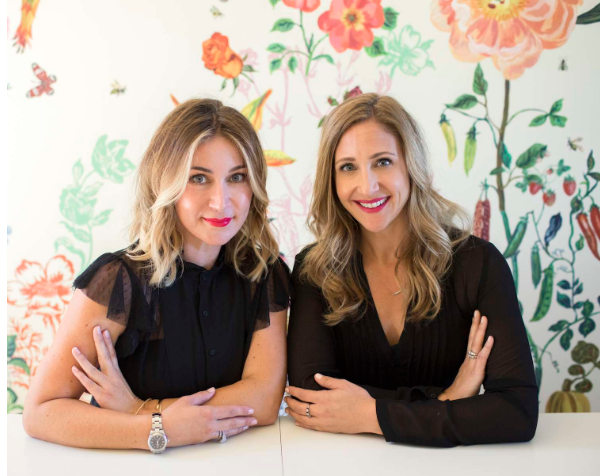 Jaime and Julie sitting with arms folded. both are blond with black shirts on. wallpaper behind them.