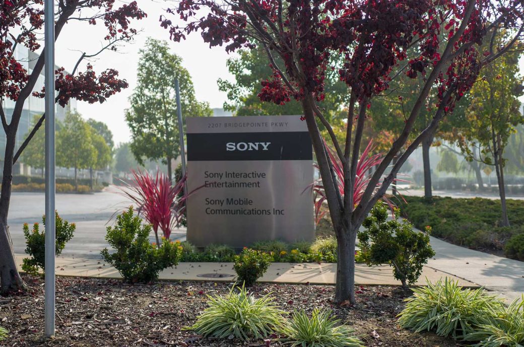 one of the many businesses in Foster City includes Sony. This is the sign for Sony Interactive Entertainment on Bridgepoint Pkwy. There are plants and a parking lot. There is a pretty tree with leaves. It looks like it must be fall.