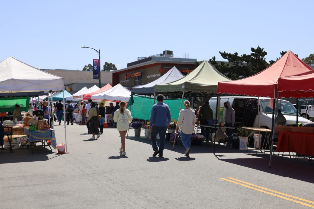 farmers market with people walking by the tents. tents are a lot of different colors.